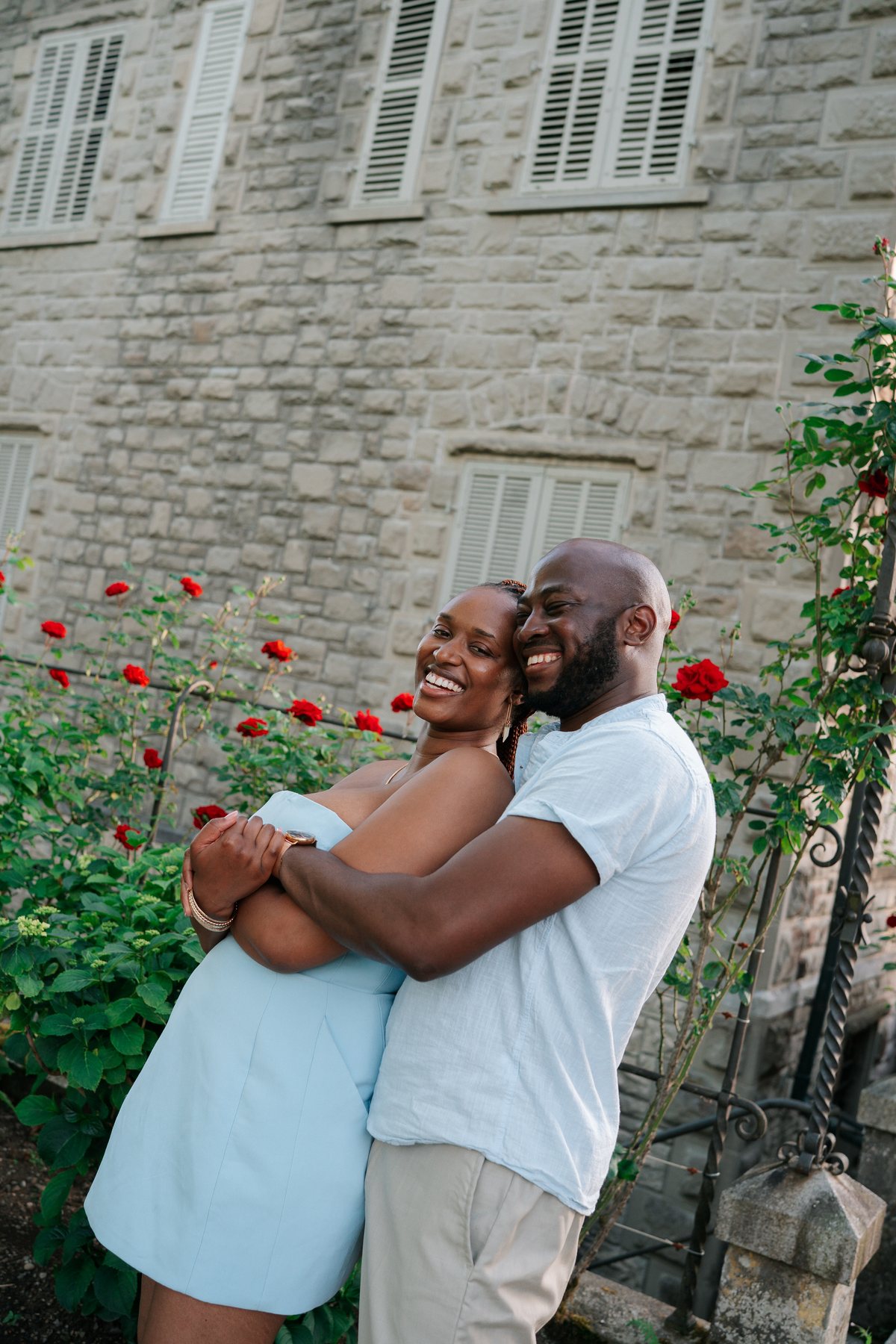 Adetunji and Sally embracing by a stone wall of red roses at golden hour